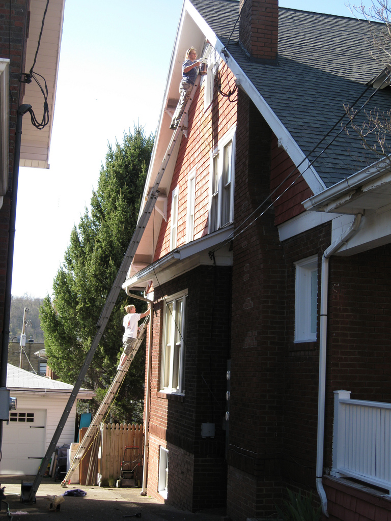 Two people on extension ladders painting the white trim of a two-story brick and cedar-shingle house.
