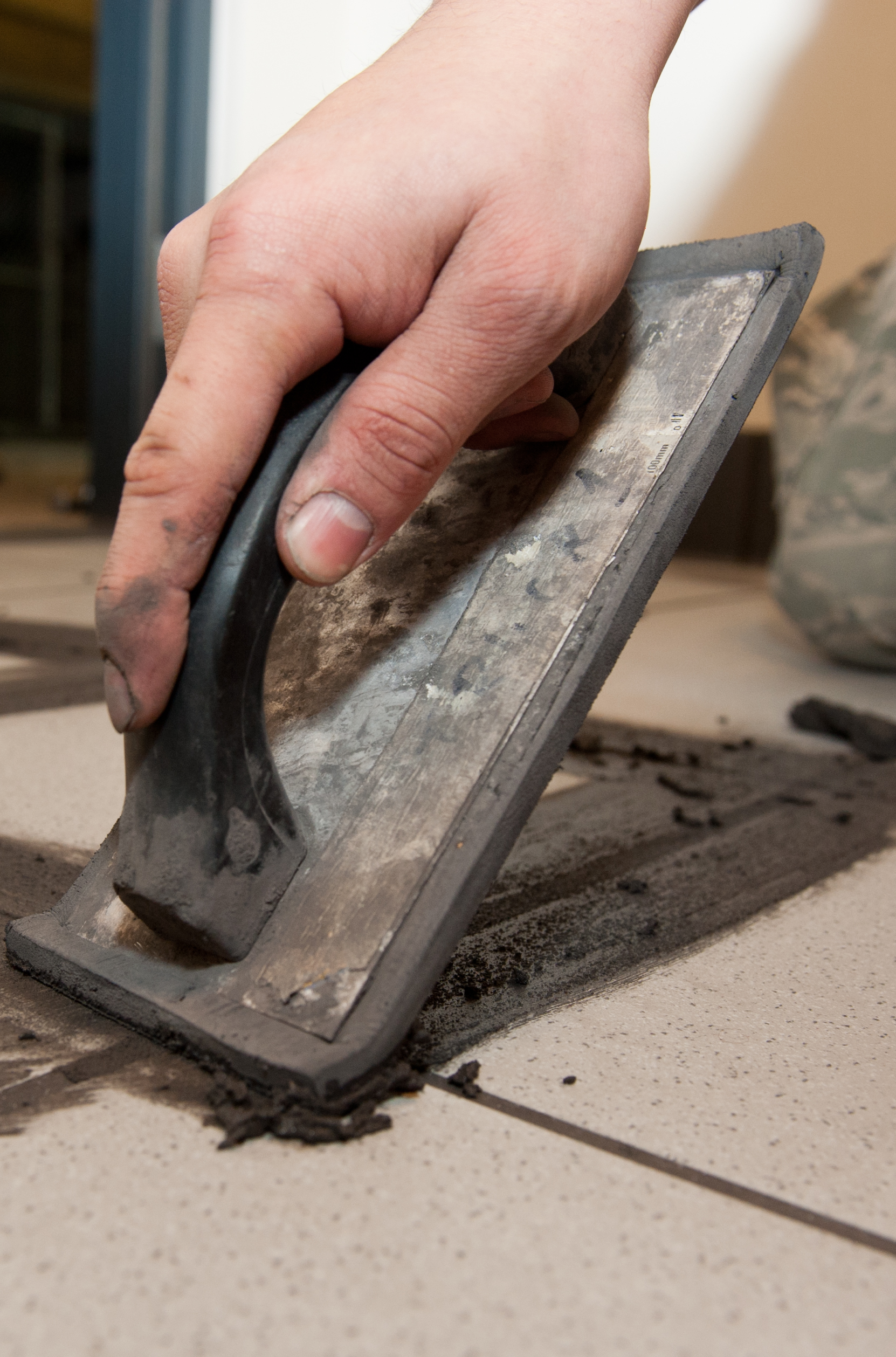 A person using a grout float to spread dark grout into the joints between light-colored floor tiles.