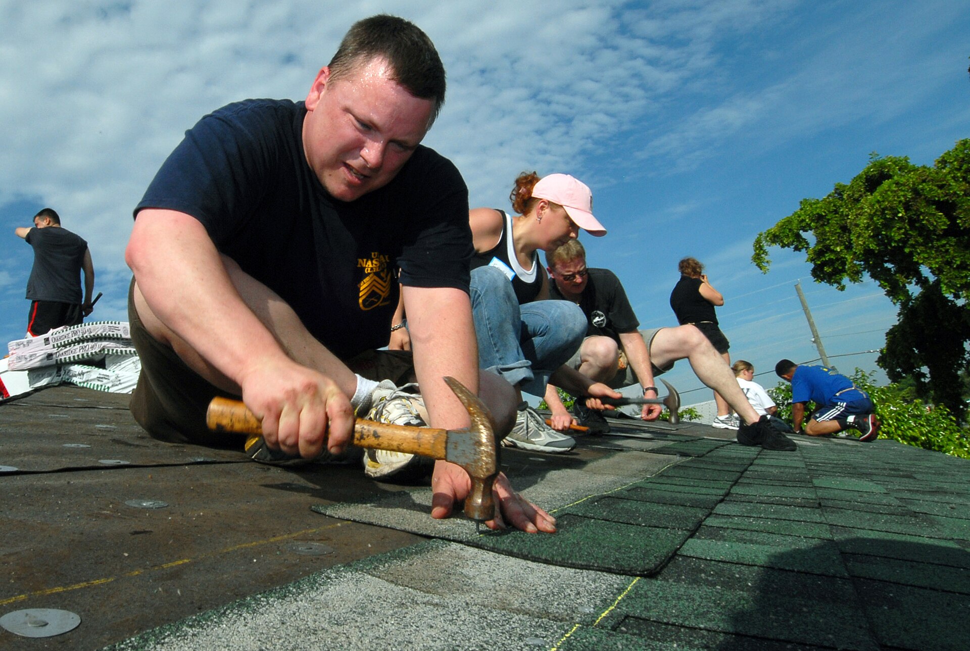 A group of people installing gray-green asphalt shingles on a sloped roof under a blue sky.